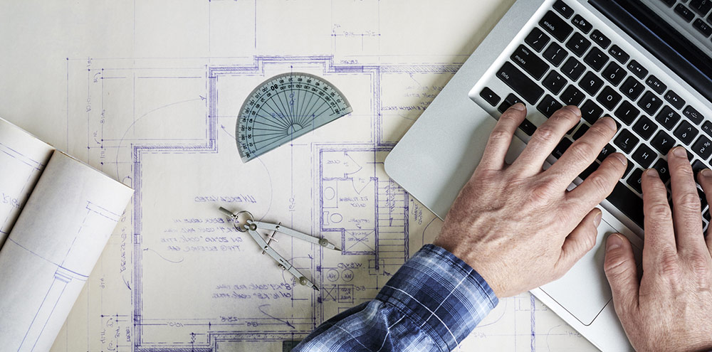 Man typing on laptop sitting on top of blueprints.